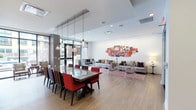 This single-floor space showcases an open-plan design that seamlessly integrates a dining area and lounge within one expansive, light-filled interior. The dining section is anchored by a substantial rectangular table with a polished stone-like surface, paired with bold red upholstered chairs and dark wood accents. Above, a row of glass pendant lights with mixed finishes adds a refined, contemporary touch to the setting.

The lounge area flows naturally from the dining space, featuring multiple seating arrangements including gray sofas accented with colorful throw pillows and sleek acrylic chairs that lend an airy, modern feel. Light hardwood flooring extends throughout, unifying the two zones while contributing to the overall warmth of the space.

A striking gallery wall composed of vibrant, red-framed artwork serves as a bold focal point on the main wall, injecting personality and visual energy into the room. Large windows and glass doors along one side allow natural light to filter in, brightening the white walls and contemporary ceiling with recessed and surface-mounted lighting. The overall ambiance is modern, lively, and welcoming.