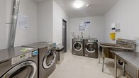 This 160 square foot utility room is a model of functional design, tailored for high-volume use in a residential or commercial setting. The space is dominated by multiple commercial-grade washing machines and dryers, indicating its primary purpose as a laundry facility. A large stainless steel sink with a deep basin is prominently featured, ideal for hand-washing or pre-treating garments. Safety and hygiene are prioritized, evidenced by the presence of a wall-mounted paper towel dispenser, hand sanitizer station, and a conspicuous yellow sharps container. The room is equipped with essential safety features, including a fire extinguisher and informational posters detailing emergency procedures. The space is well-lit with both overhead lighting and natural light from a windowed door. The floors appear to be a durable, easy-to-clean material, suitable for wet environments. Overall, the room's layout and equipment selection demonstrate a focus on efficiency, durability, and safety in a high-traffic utility area.