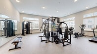 Located at 1590 Agape Way in Decatur, Georgia, this fitness center offers a well-appointed exercise space designed to support a comprehensive workout routine. The room features elevated ceilings that lend an open, airy feel, while large windows along multiple walls flood the space with abundant natural light and provide views of the surrounding exterior. The durable speckled flooring throughout is well-suited for high-traffic fitness use.

The exercise room is outfitted with a robust selection of equipment to accommodate both strength training and cardiovascular workouts. A multi-station cable and strength machine serves as the centerpiece, complemented by additional selectorized weight machines positioned throughout the room. Free weights, including a full dumbbell rack with a flat bench, provide options for varied resistance training. Cardio equipment includes treadmills, a rowing machine, and a stationary bike, ensuring a well-rounded selection for aerobic exercise.

Full-length mirrors line one wall, enhancing the sense of space while allowing users to monitor their form during workouts. Recessed lighting supplements the natural light for consistent illumination throughout the day. A wall-mounted television is also present, adding convenience for extended workout sessions. The clean, neutral color palette and thoughtfully organized layout create a focused, motivating environment for fitness.