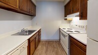 This single-floor unit offers a straightforward, functional layout across several well-defined spaces. The galley-style kitchen is efficiently arranged with rich, dark wood cabinetry lining both walls, providing ample upper and lower storage. White coil-burner appliances, including a range with overhead ventilation and a dishwasher, complement the light countertops, while wood-look vinyl flooring ties the space together with a warm, clean aesthetic.

The main living area is generously proportioned, featuring neutral carpet and light gray walls that create a calm, versatile backdrop. A sliding glass door fitted with vertical blinds introduces natural light and provides access to an outdoor view, while a secondary window further brightens the space. The transition between the entry and living zones is marked by a subtle shift in flooring materials.

Two carpeted bedrooms each offer dedicated window access, allowing natural light and exterior views. Both rooms are finished with neutral gray walls and clean white trim, providing a blank canvas adaptable to various uses.

The bathroom includes a full-size soaking tub with an integrated surround panel system featuring built-in recessed shelving on either side, paired with chrome fixtures and a showerhead above. The overall unit maintains a clean, practical design throughout.