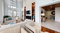 This single-floor space showcases a refined contemporary design characterized by soaring double-height ceilings and an open, flowing layout. The main lounge area centers around a striking fireplace wall, finished in sleek gray paneling with a wall-mounted flat-screen television above a linear gas fireplace. Built-in wood-toned display niches flank the feature wall, providing decorative storage highlighted by warm recessed lighting.

Natural light floods the interior through expansive floor-to-ceiling windows dressed in sheer white drapery, offering direct views of a resort-style outdoor pool and surrounding courtyard. The seating areas are furnished with plush, neutral-toned sofas and accent chairs, complemented by distinctive live-edge wood coffee tables that introduce organic warmth against the clean architectural lines.

A second zone features a reception-style counter with a coffered wood ceiling detail overhead, polished tile flooring, and a sculptural spiral staircase with black steel and metal rail detailing — adding a dramatic architectural focal point. An adjacent lounge area with wood-paneled ceilings and warm ambient lighting creates an intimate contrast to the brighter, window-lined spaces, offering a versatile and well-appointed communal environment throughout.