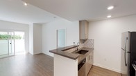 This single-level 1-bedroom, 1-bathroom residence is thoughtfully designed to maximize both functionality and style. The open-concept layout creates a seamless flow between the kitchen and living area, making the space feel airy and connected. The kitchen serves as a central feature, highlighted by a peninsula with a dark stone countertop that provides ample prep space and casual seating opportunities. Light wood cabinetry with clean-lined hardware adds warmth, while a mosaic tile backsplash introduces a subtle decorative accent. An undermount sink and built-in appliances complete the well-appointed kitchen setup. The living area extends naturally from the kitchen, offering a clean, open canvas with smooth white walls and warm wood-toned flooring that runs throughout the unit. A frosted glass door adds a modern touch while allowing light to pass between spaces. The overall palette of neutral tones — whites, warm woods, and dark stone accents — creates a contemporary and cohesive aesthetic. This residence is designed with everyday comfort in mind, offering a practical and polished environment suited to a modern lifestyle.