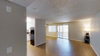 This single-floor space is designed with an open-concept layout that seamlessly connects the main living area to the kitchen. The living room is spacious and airy, featuring warm wood-style hardwood flooring that extends throughout the area, complemented by neutral-toned walls that create a clean, versatile backdrop. A textured popcorn ceiling adds a classic architectural touch overhead. Large sliding glass doors dominate one wall of the living space, flooding the interior with natural light and providing direct access to an outdoor area, while vertical blinds offer adjustable privacy and light control. A wide window adjacent to the sliding doors further enhances the bright, open atmosphere. The kitchen is partially open to the living area, separated by a structural column that defines the space without fully enclosing it. White cabinetry and countertops are visible in the kitchen, contributing to a light and functional design. Recessed and track lighting provide ample illumination throughout. The overall ambiance is practical and welcoming, with a straightforward layout that maximizes usability and flow between the main living and kitchen zones.