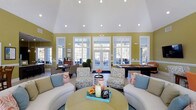This single-story clubhouse offers an inviting and well-appointed gathering space designed to foster community and comfort. The centerpiece of the space is its dramatic vaulted ceiling, which rises high above the main lounge area and is fitted with recessed lighting that illuminates the room evenly throughout the day. Warm yellow walls trimmed in crisp white molding create a cheerful, welcoming atmosphere that complements the carefully curated furnishings.

The main lounge features a mix of plush sofas and patterned accent chairs arranged around a central coffee table, providing ample seating for relaxed conversation or casual gatherings. A billiards table is positioned toward the rear of the space, adding a recreational element to the room. Adjacent seating areas offer additional spots for smaller groups to gather.

A defining feature of the clubhouse is its row of large, white-framed windows and French doors that line the back wall, flooding the interior with natural light and offering direct views and access to an outdoor patio area. The seamless connection between the indoor and outdoor spaces enhances the overall openness and airy quality of this well-designed communal facility.