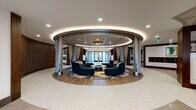 Located at 105 Corporate Park Dr in West Harrison, NY, Carraway presents a thoughtfully designed single-floor space that makes an immediate impression upon entry. The main entrance features frosted glass panel doors framed in matte black, opening onto large-format tile flooring and a welcoming mat inscribed with "Set Your Story Here," setting the tone for the experience within.

The centerpiece of the space is a dramatic circular lounge area, defined by a raised hardwood floor platform and curved rich walnut wood-paneled walls. A pair of deep teal semicircular sofas face each other around a round wood coffee table, anchored by a bold abstract area rug. Above, an elegant sculptural chandelier with cascading gold-toned branches and globe accents adds a refined artistic focal point beneath a softly illuminated tray ceiling.

Adjacent to the lounge, the reception area features a marble-clad desk with warm wood detailing, recessed lighting, and interior steel-framed windows that bring depth and transparency to the design. Nearby, a co-working or lounge table lined with deep red velvet chairs sits alongside resident mailboxes and digital display screens, offering both function and style. The overall ambiance balances contemporary sophistication with warm, residential comfort throughout.