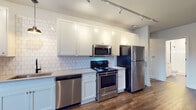 Located at 122 Parsons Ave in Columbus, Ohio, this single-floor unit offers a thoughtfully designed layout with contemporary finishes throughout. The open living area features rich, dark hardwood-style flooring and neutral-toned walls, creating a bright and welcoming atmosphere. Large windows allow natural light to fill the space while offering views of the surrounding neighborhood.

The kitchen is a standout feature, outfitted with crisp white shaker-style cabinetry, a classic white subway tile backsplash, light quartz countertops, and a full suite of stainless steel appliances including a range, microwave, dishwasher, and refrigerator. Track lighting overhead adds both function and style.

The bedrooms are carpeted for comfort and feature neutral walls and windows with blinds, providing a calm and restful environment. A well-designed hallway connects the living spaces, finished with the same dark hardwood-style flooring and white panel doors accented with matte black hardware.

The bathroom is clean and modern, showcasing floor-to-ceiling white subway tile in a walk-in shower with a glass enclosure, matte black fixtures, grey tile flooring, and a white vanity with a neutral countertop. The overall aesthetic is cohesive, combining functional design with contemporary style throughout.