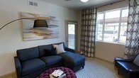 This single-floor space showcases a thoughtfully designed interior with a modern aesthetic. The living area serves as the focal point, anchored by a deep navy sectional sofa accented with decorative throw pillows, complemented by a striking tufted magenta ottoman that adds a bold pop of color. A large abstract canvas artwork in warm gold and earth tones adorns the wall, lending an artistic and sophisticated character to the room. Natural light pours in generously through a glass door and a wide window dressed with patterned geometric drapery panels, creating a bright and airy atmosphere throughout the day. The neutral-toned walls and light carpeting provide a clean, versatile backdrop that enhances the contemporary furnishings. The ceiling features a flush-mount light fixture and visible HVAC venting, reflecting practical yet unobtrusive design details. The glass door offers a glimpse of the outdoors, suggesting easy access to an exterior area. Overall, the space exudes a warm, inviting ambiance with a curated blend of bold accents and neutral tones, making it both visually engaging and comfortable for everyday living.