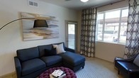 This single-floor space showcases a thoughtfully designed interior with a modern aesthetic. The living area serves as the focal point, anchored by a deep navy sectional sofa accented with decorative throw pillows, complemented by a striking tufted magenta ottoman that adds a bold pop of color. A large abstract canvas artwork in warm gold and earth tones adorns the wall, lending an artistic and sophisticated character to the room. Natural light pours in generously through a glass door and a wide window dressed with patterned geometric drapery panels, creating a bright and airy atmosphere throughout the day. The neutral-toned walls and light carpeting provide a clean, versatile backdrop that enhances the contemporary furnishings. The ceiling features a flush-mount light fixture and visible HVAC venting, reflecting practical yet unobtrusive design details. The glass door offers a glimpse of the outdoors, suggesting easy access to an exterior area. Overall, the space exudes a warm, inviting ambiance with a curated blend of bold accents and neutral tones, making it both visually engaging and comfortable for everyday living.