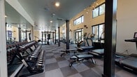 This two-story fitness center is designed to provide a comprehensive workout environment within a modern, well-appointed space. The ground floor is expansive and open, featuring soaring double-height ceilings in key areas that create an airy, energizing atmosphere. Large black-framed glass doors and clerestory windows line the perimeter, flooding the interior with natural light and offering views of surrounding palm trees and outdoor walkways. The flooring throughout consists of durable checkered rubber tiles in gray tones, providing both functionality and a clean aesthetic.

The ground level is equipped with a full dumbbell rack, adjustable weight benches, power racks with barbell storage, battle ropes, and a punching bag, offering a thorough free-weights and functional training area. Sleek black structural columns punctuate the space, complementing the industrial-modern design. Decorative sputnik-style light fixtures add a contemporary accent overhead.

A staircase with wood treads and metal railings connects to the second floor, which houses additional strength and cable machines, including multi-functional cable systems and resistance equipment. Exposed dark ceiling beams give this level a more intimate, focused atmosphere. The upper floor also features windows overlooking the surrounding area, maintaining the sense of openness consistent throughout the facility.