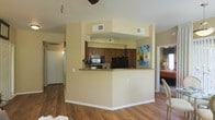 This single-floor residence showcases an open-concept design that promotes a sense of spaciousness and connectivity between the main living areas. The kitchen is a central feature, anchored by a wraparound peninsula with granite countertops that provides both workspace and casual seating. Rich, dark-toned cabinetry offers generous storage, complemented by black appliances including a refrigerator and built-in microwave. A raised soffit detail above the kitchen adds architectural definition to the space.

Warm hardwood flooring extends throughout the main living areas, creating a cohesive and inviting aesthetic. The walls are finished in a neutral, warm beige tone that enhances the brightness of the space. A glimpse into the adjacent dining area reveals additional seating and ambient lighting, while a corridor leads to further rooms beyond. The layout is practical and well-proportioned, with ceiling vents ensuring consistent climate control throughout. Colorful wall art adds a vibrant accent, contributing to the overall welcoming character of this well-designed single-level home.