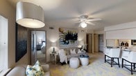 This well-appointed single-floor residence offers a thoughtfully designed layout that balances comfort and style. The living area serves as the heart of the space, featuring a ceiling fan with integrated lighting, a textured ceiling, and warm, layered ambient lighting provided by contemporary table lamps and an oversized drum pendant light. A plush sofa anchors the seating arrangement, complemented by a patterned area rug in soft yellows and whites that adds a vibrant touch to the neutral color palette. A bold, abstract wall painting serves as a striking focal point, lending an artistic character to the room. Decorative ottomans and mirrored side tables contribute to the space's modern, curated aesthetic. An open doorway provides a direct sightline into the bedroom, which is softly lit and furnished with a full bed, conveying a sense of calm and continuity throughout the unit. Adjacent to the living area, a built-in shelving unit and closet space offer practical storage solutions. The overall ambiance is warm and inviting, with a cohesive design scheme of neutral tones accented by gold and blue touches, creating a space that feels both elegant and livable.