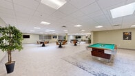 Located at 21200 Brookpark Rd in Fairview Park, Ohio, this expansive single-floor amenity space offers a generously sized, open-concept layout designed to accommodate a variety of social and recreational activities. The interior is finished with neutral-toned walls, light tile flooring, and a suspended drop ceiling with recessed panel lighting and skylights that provide bright, even illumination throughout.

The space is organized into several distinct functional zones. A full kitchen anchors one end, featuring stainless steel appliances, a large island with bar-height seating and orange accent chairs, pendant lighting, and ample cabinetry — ideal for entertaining or community events. Adjacent to the kitchen is a casual lounge area furnished with a sectional sofa, area rug, and coffee table.

The main open floor hosts multiple round dining tables with leather chairs, creating ample seating for group gatherings. A dedicated recreation zone includes a pool table and ping pong table, while a quieter lounge area offers additional seating and a wall-mounted flat-screen television. Decorative artwork is displayed throughout, adding character to the space.

Multiple access doors, generous ceiling height, and a well-planned open flow give the space a welcoming, versatile ambiance suited for both relaxed socializing and organized events.