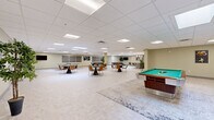 Located at 21200 Brookpark Rd in Fairview Park, Ohio, this expansive single-floor amenity space offers a generously sized, open-concept layout designed to accommodate a variety of social and recreational activities. The interior is finished with neutral-toned walls, light tile flooring, and a suspended drop ceiling with recessed panel lighting and skylights that provide bright, even illumination throughout.

The space is organized into several distinct functional zones. A full kitchen anchors one end, featuring stainless steel appliances, a large island with bar-height seating and orange accent chairs, pendant lighting, and ample cabinetry — ideal for entertaining or community events. Adjacent to the kitchen is a casual lounge area furnished with a sectional sofa, area rug, and coffee table.

The main open floor hosts multiple round dining tables with leather chairs, creating ample seating for group gatherings. A dedicated recreation zone includes a pool table and ping pong table, while a quieter lounge area offers additional seating and a wall-mounted flat-screen television. Decorative artwork is displayed throughout, adding character to the space.

Multiple access doors, generous ceiling height, and a well-planned open flow give the space a welcoming, versatile ambiance suited for both relaxed socializing and organized events.