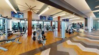 This expansive fitness center is thoughtfully designed to accommodate a wide range of workout styles and fitness goals. The space is organized into several distinct zones, beginning with a welcoming entry corridor that immediately sets the tone with a bold, eye-catching geometric wall mural in cool icy blues and whites. Underfoot, a striking large-scale chevron-patterned floor in mixed tones of wood, gray, and charcoal runs throughout much of the facility, adding a dynamic visual element to the overall design.

The main strength and conditioning area features an extensive selection of cable machines, free weights, and specialty equipment, anchored by warm wood-toned cylindrical columns and oversized ceiling fans. Mounted flat-screen TVs keep members entertained during workouts, while floor-to-ceiling windows flood the space with abundant natural light and offer open views of the surrounding outdoor areas.

A dedicated cardio zone lines the perimeter with rows of treadmills and other aerobic machines positioned directly in front of expansive glass walls, maximizing the connection to the outdoors. An additional area accommodates rowing machines and stair climbers.

A separate spin studio provides a focused, high-energy environment, furnished with multiple stationary bikes, full-wall mirrors, and teal-accented walls that complement the broader color palette of the facility. The overall ambiance is modern, energetic, and polished.