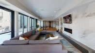 Located at 1300 W Lake St in Minneapolis, Minnesota, this single-floor amenity lounge is a thoughtfully designed communal space that blends modern aesthetics with inviting functionality. Rich hardwood flooring flows throughout the open-plan layout, connecting distinct activity zones in a seamless, cohesive manner. The centerpiece of the main lounge area is a dramatic floor-to-ceiling marble-clad accent wall, framing a sleek linear gas fireplace and a wall-mounted television — creating a warm, sophisticated focal point. Generous sectional seating is arranged to encourage relaxed gathering, while abundant recessed lighting complements the natural light that pours in through an expansive wall of floor-to-ceiling sliding glass doors. These doors open directly onto an outdoor terrace, offering views of the surrounding Minneapolis cityscape. A dedicated game area features a billiards table, adding a recreational element to the space. The kitchenette is appointed with rich walnut cabinetry, stone countertops, a marble backsplash, and a built-in beverage cooler, making it well-suited for entertaining. A semi-private meeting or dining nook with a round table and mid-century-inspired chairs offers a more intimate setting, enhanced by a stylish globe floor lamp. Curated artwork and lush potted plants are featured throughout, lending the space a gallery-like, welcoming ambiance.