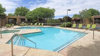Located at 4401 Morris St NE in Albuquerque, New Mexico, this outdoor amenity area is centered around a large, well-maintained swimming pool with clear turquoise water. The pool deck is finished in natural stone-look pavers, providing a generous sunbathing and lounging area equipped with multiple green sling-style chaise lounges and chairs arranged for comfort and relaxation.

Adjacent to the pool is a covered pergola structure attached to the main building, which features warm earth-toned stucco exteriors characteristic of Southwestern architectural style. The pergola area is furnished with additional seating, making it an ideal shaded retreat on sunny New Mexico days. The surrounding buildings display a blend of deep red, tan, and brown tones, complementing the desert aesthetic of the region.

The pool area is fully enclosed by a metal fence, ensuring safety and privacy, with gated access points. Lush mature trees and well-maintained shrubs and hedges border the perimeter, adding greenery and a sense of seclusion. Accent lighting poles are positioned throughout the deck, allowing the space to be enjoyed into the evening hours. The overall ambiance is inviting and resort-like, offering a comfortable outdoor community space suited to the Albuquerque climate.