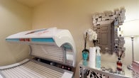 This single-level space is designed as a dedicated tanning room, centered around a professional-grade Ergoline tanning bed that serves as the focal point of the room. The unit features a streamlined white and teal design with built-in UV lighting, offering a high-end tanning experience. The walls are finished in warm neutral tones, creating a calm and welcoming ambiance suited to a personal care environment. Adjacent to the tanning bed is a mirrored glass side table, neatly arranged with tanning lotions, sanitizing sprays, and paper towels, ensuring all essentials are within reach. A decorative mosaic-style mirror mounted on the wall adds a modern, polished touch to the space, complementing the metallic accents throughout. A small floral arrangement featuring white orchids in a teal vase introduces a subtle natural element, softening the overall aesthetic. The room's compact, focused layout makes efficient use of the available space, ensuring a clean, organized, and professional atmosphere ideal for a personal or commercial tanning service setting.