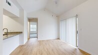This single-floor, two-bedroom, one-bathroom unit makes efficient use of its compact footprint through a thoughtful layout. The living room is an immediate standout, featuring striking vaulted ceilings that amplify the sense of vertical space and openness well beyond what the floor area alone might suggest. Light hardwood flooring runs throughout, contributing to a bright, clean aesthetic that complements the crisp white walls. A pendant light fixture hangs from the peak of the vaulted ceiling, adding a subtle design detail. A large sliding door dressed with vertical blinds allows for natural light while offering privacy control. The primary bedroom, accessible through the hallway, is a comfortable size and benefits from its own window, bringing in additional natural light. A second bedroom provides flexibility for guests or a home office. The unit notably includes two kitchen areas, offering practical versatility for cooking and storage. The single bathroom and connecting hallway complete the efficient floor plan. Overall, the unit delivers a bright, airy ambiance anchored by its dramatic ceiling height and neutral, light-toned finishes throughout.
