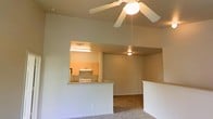 This single-story space features an open-concept layout that connects the living area to the kitchen through a functional pass-through counter, enhancing the sense of openness and flow throughout the unit. The living area is finished with wall-to-wall carpeting and is anchored by a ceiling fan with a built-in light fixture, which provides both air circulation and ambient lighting. The ceiling height is notably generous, contributing to an airy and spacious feel.

The kitchen is visible through the pass-through opening and is equipped with light wood cabinetry, a range, and overhead fluorescent lighting that illuminates the workspace effectively. A secondary room or hallway is visible beyond the main living space, suggesting additional functional areas within the layout. The neutral wall tones and light color palette throughout create a clean, understated aesthetic that offers versatility for furnishing and decorating. The pass-through counter also functions as a breakfast bar-style surface, providing a casual dining or serving option that bridges the kitchen and living areas.