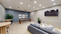 Located in Aurora, CO, this well-appointed community lounge offers a thoughtfully designed single-floor layout that balances comfort and entertainment. The living area serves as the focal point of the space, anchored by a plush gray sectional sofa resting on a gold-toned patterned area rug over light wood-look flooring that runs throughout the room. A wall-mounted flat-screen TV faces the seating arrangement, while a cluster of modern accent tables completes the lounging setup.

A striking slate-blue accent wall adds depth and contrast to the otherwise warm, neutral palette of greiges and whites. Pendant lighting hangs above a wood shuffleboard table positioned along the accent wall, which is further adorned with a trio of framed landscape photographs. Bar-height round tables with sleek stools provide additional social seating in the corner, accompanied by lush potted plants that introduce a touch of greenery. Recessed ceiling lighting illuminates the entire space evenly, creating a bright and welcoming ambiance. A frosted-glass door provides a stylish entry point, completing this versatile and inviting communal space.