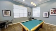 This inviting game room is designed for leisure and entertainment, centered around a full-size pool table with a teal felt surface and classic wooden frame. The room is finished with soft blue-gray walls, crisp white crown molding, and white baseboards that lend a clean, polished aesthetic throughout. Large windows dressed with white horizontal blinds fill the space with generous natural light, creating a bright and welcoming atmosphere. Overhead, a stylish pendant light fixture with multiple frosted glass shades hangs above the pool table, providing focused task lighting that complements the room's overall ambiance. In the background, a small bistro-style table with black chairs offers an intimate seating area, ideal for spectators or casual conversation. Framed artwork on the walls adds a decorative touch, reinforcing the room's thoughtfully curated design. The flooring features a neutral stone or tile pattern that grounds the space with a durable, easy-to-maintain surface. Together, these elements create a versatile and functional recreation space that balances style with practicality, making it well-suited for both casual play and social gatherings.