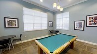 This inviting game room is designed for leisure and entertainment, centered around a full-size pool table with a teal felt surface and classic wooden frame. The room is finished with soft blue-gray walls, crisp white crown molding, and white baseboards that lend a clean, polished aesthetic throughout. Large windows dressed with white horizontal blinds fill the space with generous natural light, creating a bright and welcoming atmosphere. Overhead, a stylish pendant light fixture with multiple frosted glass shades hangs above the pool table, providing focused task lighting that complements the room's overall ambiance. In the background, a small bistro-style table with black chairs offers an intimate seating area, ideal for spectators or casual conversation. Framed artwork on the walls adds a decorative touch, reinforcing the room's thoughtfully curated design. The flooring features a neutral stone or tile pattern that grounds the space with a durable, easy-to-maintain surface. Together, these elements create a versatile and functional recreation space that balances style with practicality, making it well-suited for both casual play and social gatherings.