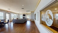 This single-story community facility is thoughtfully designed to offer both comfort and functionality across several distinct areas. The space is unified by rich hardwood-style flooring, recessed lighting, and a coastal-inspired color palette of soft blues, whites, and warm gold accents reflected in the crown molding and wall trim details throughout.

The reception area greets visitors with an elegantly curved front desk finished in white paneling with a dark stone countertop. Elevated ceilings with gold-trimmed molding create a sense of openness, while framed coastal artwork and direct exterior access contribute to a welcoming atmosphere.

The spacious lounge area serves as the heart of the facility, featuring generous sectional and sofa seating in neutral gray tones, accented with blue and earth-toned throw pillows. A built-in media unit with an arched surround houses a flat-screen television, flanked by multiple windows that flood the room with natural light and offer views of the surrounding greenery. Decorative framed windows and wall-mounted artwork add architectural interest throughout.

The fitness room is compact yet well-equipped, featuring professional-grade treadmills and a cross-trainer positioned to take advantage of natural light from large windows overlooking mature landscaping. A wall-mounted television and safety mirror complete the space, providing a functional and bright environment for exercise.
