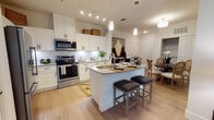 This single-floor residence showcases a thoughtfully designed open-concept layout that seamlessly connects the kitchen, dining, and living areas. The kitchen is a standout feature, outfitted with crisp white shaker cabinets, a geometric hexagonal tile backsplash, stainless steel appliances including a range, microwave, and bottom-freezer refrigerator, and light quartz countertops. A central island with an undermount sink and seating for two provides both functionality and a natural gathering point. Track lighting and elegant pendant lights illuminate the space, while light-toned hardwood-style flooring flows throughout the main living areas. The dining area offers comfortable seating adjacent to the kitchen, and the living room features large floor-to-ceiling curtained windows that invite natural light and create an airy atmosphere. The bedroom is carpeted for warmth and comfort, with windows fitted with blinds and sheer curtains providing ample natural light. The bathroom presents a clean, modern aesthetic with a quartz vanity countertop, undermount sink, wide mirror, white cabinetry, and a tiled shower-tub combination. Throughout the residence, neutral wall tones and consistent finishes create a cohesive, contemporary ambiance well-suited for comfortable everyday living.