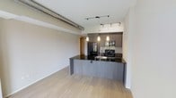This single-floor space showcases a sleek, modern design with an open-concept layout that seamlessly connects the kitchen and living area. The kitchen is a standout feature, equipped with dark, flat-panel cabinetry, a striking black granite countertop, and a peninsula that doubles as a casual dining or prep space. Black stainless steel appliances, including a refrigerator and gas range, complement the contemporary aesthetic, while a tasteful tile backsplash adds a refined touch. Pendant lighting and a track lighting system illuminate the kitchen area with style and precision. Light-toned hardwood flooring runs throughout the space, providing a warm contrast to the darker cabinetry and finishes. An exposed cylindrical ventilation duct runs along the ceiling, lending an industrial character to the otherwise polished interior. The open living area offers a generous, flexible space bathed in a neutral palette, creating a bright and airy ambiance. A hallway visible in the background suggests additional private rooms beyond the main living area. Overall, the space strikes a balance between industrial and contemporary design, offering a functional and visually cohesive environment.