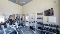 Located in Dallas, this fitness center offers a comprehensive workout environment designed to accommodate a range of exercise needs. The space features soaring ceilings that create an open, airy atmosphere, complemented by clean white walls and neutral-toned flooring with a textured, dark finish that adds both style and durability underfoot. Large mirrored panels line one wall, visually expanding the space while allowing users to monitor their form during workouts. The facility is outfitted with a variety of cardio equipment, including a treadmill and a dedicated spin bike, ideal for cardiovascular training. A multi-functional cable weight machine anchors the strength training area, providing versatility for resistance exercises. A well-organized dumbbell rack along the far wall offers a full range of free weights for targeted strength work. A wall-mounted television provides entertainment during workouts. Ventilation is supported by ceiling vents and a ceiling fan, helping to maintain a comfortable environment. The overall ambiance is clean, modern, and functional, making this fitness center a practical and well-appointed space for daily exercise.