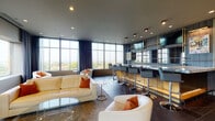 Located at 797 N Wall St in the heart of Columbus, Ohio, this elevated lounge space delivers a sleek, contemporary atmosphere defined by clean lines and upscale finishes. The main lounge area is anchored by a striking full-service bar setup along one wall, complete with a light wood-paneled front, dark stone countertop, and a row of modern high-back bar stools in charcoal gray. Above the bar, open metal shelving with gold accents and mounted flat-screen televisions add a polished, entertainment-ready aesthetic. Opposite the bar, a generously sized seating arrangement features curved white leather sofas accented with bold orange throw pillows, surrounding a glass-top coffee table — creating an inviting social hub. The space is bathed in natural light thanks to expansive floor-to-ceiling windows that wrap around a corner, offering panoramic views of the Columbus skyline and surrounding treetops. Dark charcoal tile flooring and a matching ceiling with recessed lighting complete the refined, contemporary look. The overall ambiance is that of a high-end rooftop lounge — sophisticated, functional, and ideal for both relaxed gatherings and more formal entertaining.