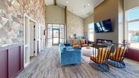 Located in Box Elder, South Dakota, this single-story community clubhouse at 10Bedrock offers a thoughtfully designed gathering space that blends modern aesthetics with warm, inviting elements. The main lounge area makes an immediate impression with its soaring vaulted ceilings accented by exposed wood beams, a dramatic floor-to-ceiling stacked stone accent wall, and large windows that flood the space with natural light. Comfortable seating, including a teal velvet sofa and cognac leather accent chairs, is arranged around a sleek linear fireplace, creating an ideal environment for relaxation and socializing. A built-in refreshment bar with cabinetry, a mini-fridge, and a coffee station adds everyday convenience. The adjacent kitchen is well-appointed with gray shaker-style cabinetry, stainless steel appliances including a full-size refrigerator, a center island with bar seating, pendant lighting, and light hardwood-style flooring — all complemented by a striking mosaic-tiled feature wall. A secondary lounge area features rich plum wainscoting, wall-mounted media, and flexible seating, suitable for meetings or quiet use. Throughout the space, layered textures, decorative tile work, and curated furnishings come together to create a polished yet approachable community amenity.