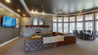This well-appointed single-floor communal space offers a blend of rustic charm and modern comfort across several functional areas. The centerpiece of the main lounge is a striking full-height natural stone fireplace wall, complete with a substantial wood mantel and arched hearth, creating a warm and inviting focal point. Generous tufted leather sectional seating surrounds a glass coffee table, ideal for relaxed gatherings. Large curved banks of floor-to-ceiling windows flood the space with natural light and provide views of the surrounding landscape, enhancing the open and airy atmosphere. Crown molding and recessed lighting throughout add a refined architectural touch, while the dark painted ceilings lend a sophisticated contrast to the lighter walls. An adjacent dining area accommodates a formal table and chairs, positioned near additional oversized windows and a glass-panel door leading outside. A compact but fully equipped kitchen features stainless steel appliances, white cabinetry, and a classic white subway tile backsplash, providing practical amenities for events and gatherings. Decorative columns and wide corridors connect the various zones seamlessly, giving the space a cohesive and polished feel throughout.