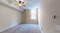 Located in Cincinnati's 45202 zip code, this single-floor residence at 207 Eighth St E offers a thoughtfully designed layout with a neutral, move-in-ready aesthetic throughout. The kitchen serves as a functional centerpiece, outfitted with rich wood cabinetry, granite countertops, a subway tile backsplash, and a full suite of stainless steel appliances including a French door refrigerator, gas range with microwave, and a double-basin sink. A central island provides additional prep and gathering space, while tile flooring adds durability underfoot. The living and sleeping areas feature plush wall-to-wall carpeting in a soft gray tone, complemented by warm beige walls and clean white trim. Ceiling fans are present in the main carpeted rooms, promoting airflow and comfort. Natural light enters through tall, double-hung windows that frame views of the surrounding brick urban streetscape, lending an airy quality to the interior. The bathroom is finished with light tile flooring, a warm wood vanity with ample cabinetry and drawers, a white countertop, and a framed mirror with overhead lighting. The overall ambiance is clean, understated, and well-maintained, offering a comfortable urban living environment in the heart of Cincinnati.