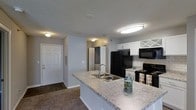 This single-story residence on Lake Lynn Drive in Raleigh, NC, offers a practical and well-appointed layout designed for everyday comfort. The kitchen serves as a central highlight, featuring a spacious granite-style countertop peninsula with a double stainless steel sink, ideal for both meal preparation and casual dining. White cabinetry with decorative glass-front upper panels lines the walls, complemented by a classic subway tile backsplash that adds a clean, timeless aesthetic. Black appliances, including a refrigerator, electric range, and over-the-range microwave, provide a bold contrast against the lighter cabinetry and countertops. Flush-mount ceiling lighting keeps the space bright and functional. The kitchen opens directly toward the entryway, where a standard front door with an adjacent utility area is visible, contributing to the home's efficient flow. Carpeted flooring transitions to hard surface flooring near the entry. A textured ceiling finish is consistent throughout the visible areas. The overall ambiance is neutral and move-in ready, with a clean, understated palette suited to a variety of interior styles.