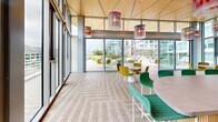 Located at 222 N City Dr in San Marcos, CA, this thoughtfully designed amenity space seamlessly blends an interior lounge with an expansive outdoor terrace. The interior is anchored by soaring floor-to-ceiling glass walls that flood the space with natural light and offer unobstructed views of the surrounding urban landscape. A warm wood-paneled ceiling adds an organic contrast to the modern architecture, while artistic pendant light fixtures in layered, translucent designs serve as striking focal points throughout. The lounge features a built-in bar with woven teal counter stools, a large-format display screen set against a richly textured, multi-toned feature wall, and an eclectic mix of woven seating in bold yellow and green hues arranged around contemporary tables. The patterned herringbone flooring adds visual depth and texture underfoot. The space opens directly onto a generously sized outdoor terrace, where raised planters filled with ornamental grasses and native landscaping create a serene, garden-like atmosphere. A professional outdoor kitchen outfitted with multiple stainless steel grills provides an exceptional entertaining amenity. Lounge seating is scattered across the paver-tiled terrace, offering open-air views of the neighboring mixed-use development. Together, the indoor and outdoor areas create a cohesive, resort-inspired environment ideal for gathering and relaxation.
