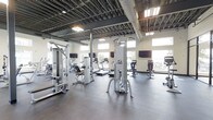 This fitness center offers a spacious, open-plan workout environment designed to accommodate a full range of exercise needs. The facility is outfitted with professional-grade Cybex strength training machines, including cable systems, seated resistance stations, and functional training towers, thoughtfully arranged to allow ample room for movement between equipment. Cardio machines, including treadmills, are positioned along the windowed perimeter, allowing exercisers to enjoy natural light and outdoor views during their workouts. Multiple wall-mounted flat-screen televisions are installed throughout the space, providing entertainment and engagement for users across different workout zones. The flooring consists of durable dark rubber matting, ideal for high-traffic fitness use and providing safety and comfort underfoot. The ceiling is left exposed, showcasing the building's structural framework and ductwork, lending the space an industrial aesthetic that complements its modern fitness equipment. Large windows along the walls invite abundant natural light, creating an energizing and open atmosphere. The overall design balances functionality with a clean, modern aesthetic, making it a well-appointed facility suited for both strength and cardio training routines.