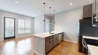 Located at Grandview Crossing in Columbus, Ohio, this two-bedroom, two-bathroom residence offers a well-designed single-floor layout with modern finishes throughout. The open-concept kitchen and living area serves as the heart of the home, anchored by a large center island with a white quartz countertop, undermount sink, and matte black pendant lighting above. Gray cabinetry pairs with stainless steel appliances, a subway tile backsplash, and warm hardwood-style flooring to create a clean, contemporary aesthetic. Natural light flows generously through large windows dressed with horizontal blinds. The living area extends seamlessly from the kitchen, providing an airy, connected feel. The primary bedroom features hardwood-style flooring, large windows with outdoor views, and direct access to its en-suite bathroom, which includes a walk-in tiled shower, a gray vanity with white countertop, matte black fixtures, and large-format tile flooring. The second bedroom is carpeted and comfortably proportioned, served by a second full bathroom with a soaking tub and matching contemporary finishes. Both closets offer built-in shelving for organized storage. A hallway connects all rooms efficiently, completing this well-appointed residence.