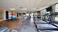 Located at University Hills Blvd in Toledo, Ohio, this fitness center offers a comprehensive workout environment within a generously sized, single-floor layout. The space is designed to accommodate a wide range of exercise needs, with clearly defined zones for both cardio and strength training.

The cardio area features a row of treadmills and elliptical machines positioned along large windows, providing natural light and outdoor views of surrounding greenery—an inviting backdrop during workouts. Wall-mounted televisions are positioned throughout the space, keeping users entertained during extended sessions.

The strength training section is well-stocked with cable machines, multi-use weight stations, adjustable benches, and free weights, offering versatile options for resistance training. A dedicated functional fitness area includes a rack of exercise balls in various sizes, a heavy bag, and open floor space suitable for stretching or dynamic movement.

Full-length mirrors line one wall, enhancing the sense of openness and allowing users to monitor their form. The flooring throughout is durable rubber, ideal for a high-traffic fitness environment. Recessed drop-panel ceilings with integrated lighting provide consistent, even illumination across the entire space. The overall atmosphere is clean, functional, and purpose-built for an effective fitness experience.