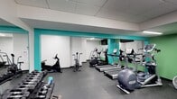 This fitness center is designed to support a well-rounded workout experience within a single-floor layout. The space is furnished with a selection of modern cardio machines, including treadmills, ellipticals, and stationary bikes from recognized brands such as TRUE and Precor, arranged to allow comfortable movement between stations. A dedicated free weights area lines one side of the room, offering a range of dumbbells for strength training. The flooring consists of durable rubber tiles, providing grip and shock absorption suited to high-activity use. Walls are finished in white with bold teal accent bands that lend a clean, energetic aesthetic to the interior. A large mirrored wall along one side visually expands the space while allowing users to monitor their form during exercise. A wall-mounted flat-screen television is positioned for visibility from the cardio equipment. The drop ceiling with recessed lighting ensures consistent, even illumination throughout. Overall, the space maintains a practical and modern atmosphere, thoughtfully outfitted to meet a variety of fitness needs.