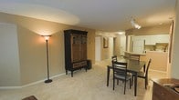 This single-floor residence offers a comfortable and functional layout centered around an open-plan living and dining area. The dining space is defined by a dark-toned table and chair set, complemented by warm beige carpeted flooring that extends throughout the main living areas, contributing to a cozy and inviting atmosphere. A dark wood armoire with glass-panel upper doors serves as a notable storage piece and adds a classic character to the room. Warm ambient lighting from a floor lamp enhances the neutral-toned walls, creating a welcoming environment throughout. Track lighting overhead provides additional illumination to the open-plan area. A hallway extends from the main living space, connecting to additional rooms and featuring a framed wall accent, adding a touch of décor to the transitional area. Adjacent to the dining area, a kitchen is visible and includes a raised countertop overhang that functions as a breakfast bar, with seating and overhead cabinetry. The space is practical and well-organized, with a single-floor plan that ensures ease of movement between the living, dining, and kitchen areas. The overall ambiance is warm and understated, suited to comfortable everyday living.