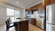 This well-appointed single-floor residence offers a thoughtfully designed layout that balances modern aesthetics with functional living. The kitchen serves as a standout centerpiece, featuring rich walnut-toned cabinetry extending to the ceiling, a herringbone-patterned tile backsplash, quartz countertops, and a full suite of stainless steel appliances including a range, over-the-range microwave, dishwasher, and refrigerator. A large central island with bar seating provides additional prep space and a casual dining option, while light hardwood flooring ties the kitchen seamlessly into the adjoining living area. The living room is bright and welcoming, with large windows that invite abundant natural light and offer views of the surrounding neighborhood. The two bedrooms are carpeted for comfort, each with access to natural light; the primary bedroom features sliding glass doors opening to a balcony with outdoor views. Two bathrooms echo the home's modern design language, showcasing walnut-finish vanities with white quartz countertops, large-format tile surrounds in the tub and shower areas, and wood-look flooring. Clean lines, neutral tones, and quality finishes are consistent throughout, creating a cohesive and contemporary living environment.
