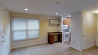 This single-floor space offers a functional and straightforward layout designed to maximize utility. The main living area features neutral beige carpet flooring and light-toned walls that create a bright, understated atmosphere. Recessed ceiling lights provide consistent overhead illumination, supplemented by natural light streaming through a sizable window fitted with horizontal blinds. A wall-mounted air conditioning unit serves the space for climate control.

A notable built-in feature is the wood-toned desk with a granite-style surface and integrated drawer storage, positioned along the interior wall — ideal for a dedicated workspace. The carpeted living area transitions to hard-surface flooring as it opens into the adjacent kitchen.

The kitchen, visible through a wide pass-through opening, is efficiently arranged with medium-toned wood cabinetry providing ample storage above. White appliances, including an electric range and an over-the-range microwave, lend a clean, classic appearance. The overall layout is practical and compact, making it well-suited for individuals seeking a low-maintenance, single-level living arrangement with essential amenities in close proximity.