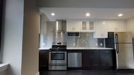 This single-floor space showcases a thoughtfully designed interior that balances modern aesthetics with functional living. The kitchen is a standout feature, outfitted with a full suite of stainless steel appliances, including a gas range, built-in microwave, and dishwasher. Dark espresso lower cabinets are paired with crisp white upper cabinetry, creating a striking two-tone contrast. A metallic mosaic tile backsplash adds texture and visual interest, while a prominent stainless steel wall-mounted range hood serves as a focal point above the cooktop. Light-colored countertops and recessed lighting complete the polished look. The living area transitions seamlessly from the kitchen, featuring rich dark hardwood flooring that runs throughout the space, adding warmth and depth. Neutral-toned walls provide a clean, versatile backdrop, while large windows with dark frames flood the room with natural light and frame elevated city views, offering a strong sense of openness and connection to the urban landscape. The architectural detailing includes prominent white baseboards and structural column accents that lend a refined character to the space. Overall, the unit presents a sleek, contemporary atmosphere well-suited for modern urban living.