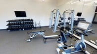 This fitness center is designed to support a full range of workout routines in a clean, organized environment. The space features durable rubber flooring throughout, providing both safety and comfort during exercise. A full-length wall mirror enhances the sense of space while allowing users to monitor their form during training sessions.

The strength training area is well-appointed, featuring a multi-station cable machine ideal for a variety of resistance exercises. An adjustable workout bench offers versatility for both flat and incline movements. A complete dumbbell rack lines one wall, stocked with an extensive range of weights to accommodate all fitness levels. A Matrix-branded piece of equipment adds to the cardio and functional training options available.

The space is finished with bright, neutral walls and efficient overhead lighting that keeps the environment well-lit and energizing. A wall-mounted flat-screen television is positioned to keep users entertained during their workouts. The overall layout is thoughtfully organized to maximize usable floor space while ensuring safe clearance between equipment. This fitness center provides everything needed for a comprehensive and effective workout experience.