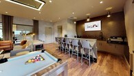 The Club Lounge at 1177 Warburton Ave in Yonkers, NY is a thoughtfully designed amenity space that seamlessly blends entertainment, relaxation, and productivity. The open-plan layout is anchored by warm hardwood flooring in a herringbone-inspired pattern, textured concrete-finish walls, and recessed lighting that creates a welcoming, upscale ambiance throughout.

The lounge area features a plush gray tufted sofa and cognac leather armchairs arranged around a sleek white coffee table, complemented by a wall-mounted flat-screen TV and a wooden media console. Adjacent to the seating zone, a professional billiards table with modern chrome legs takes center stage beneath a rectangular pendant light fixture, adding a focal point to the entertainment experience.

The wet bar area is finished with a richly textured, dark decorative tile backsplash, a wall-mounted television, under-counter cabinetry, and a sink — all set against a long counter lined with stylish bar stools. A second bar-height counter with additional seating provides a versatile space for dining or socializing.

A dedicated workstation nook houses dual computer monitors set along a sleek desk positioned beneath large windows, flooding the area with natural light and views of the surrounding greenery. A glass-enclosed room nearby offers additional private space. The overall design is cohesive and contemporary, making this lounge a well-rounded community hub.