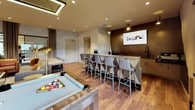 The Club Lounge at 1177 Warburton Ave in Yonkers, NY is a thoughtfully designed amenity space that seamlessly blends entertainment, relaxation, and productivity. The open-plan layout is anchored by warm hardwood flooring in a herringbone-inspired pattern, textured concrete-finish walls, and recessed lighting that creates a welcoming, upscale ambiance throughout.

The lounge area features a plush gray tufted sofa and cognac leather armchairs arranged around a sleek white coffee table, complemented by a wall-mounted flat-screen TV and a wooden media console. Adjacent to the seating zone, a professional billiards table with modern chrome legs takes center stage beneath a rectangular pendant light fixture, adding a focal point to the entertainment experience.

The wet bar area is finished with a richly textured, dark decorative tile backsplash, a wall-mounted television, under-counter cabinetry, and a sink — all set against a long counter lined with stylish bar stools. A second bar-height counter with additional seating provides a versatile space for dining or socializing.

A dedicated workstation nook houses dual computer monitors set along a sleek desk positioned beneath large windows, flooding the area with natural light and views of the surrounding greenery. A glass-enclosed room nearby offers additional private space. The overall design is cohesive and contemporary, making this lounge a well-rounded community hub.