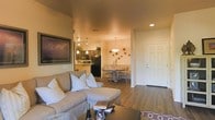 This single-story residence showcases an open-concept layout that seamlessly connects the living room, dining area, and kitchen into one cohesive, flowing space. The living room is anchored by a plush, neutral-toned sectional sofa accented with decorative pillows, paired with a dark wood coffee table that grounds the seating area. A large framed wall art piece featuring a dramatic landscape adds a bold focal point to the room. Warm recessed lighting and pendant fixtures cast an inviting amber glow throughout the space, enhancing the rich, hardwood-style flooring that runs consistently across all areas. The kitchen, visible from the living room, features dark cabinetry, stainless steel and black appliances, and a breakfast bar with pendant lighting overhead. A dedicated dining area sits adjacent, furnished with a round table and upholstered chairs beneath an elegant hanging light fixture. Subtle wall décor, including scattered circular ornaments, adds character to the dining space. A white paneled entry door provides a clean, classic contrast to the warm interior palette. The overall ambiance is cozy, modern, and well-appointed.
