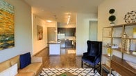 This single-floor residence showcases a thoughtfully designed open-concept layout that seamlessly connects the living and kitchen areas, creating a spacious and cohesive flow. Rich hardwood flooring runs throughout, grounding the space with warmth and continuity.

The kitchen is a standout feature, outfitted with dark espresso cabinetry, stainless steel appliances, and a breakfast bar peninsula that doubles as a casual dining and prep space. A track lighting system overhead provides focused illumination, complementing the pendant lighting above the peninsula for a layered, modern aesthetic. Light-toned walls contrast beautifully with the darker cabinetry, keeping the space feeling bright and open.

The living area is comfortably proportioned, with room for a full seating arrangement. A bold piece of wall art adds a touch of personality, while a geometric-patterned area rug anchors the space beneath the furnishings. Clean architectural lines, including defined ceiling transitions and efficient use of vertical space, contribute to the contemporary character of the residence.

Overall, the space strikes a balance between modern functionality and warm, livable design — making it well-suited for both everyday living and casual entertaining.