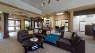 This single-story community clubhouse is designed to offer residents a comfortable and functional gathering space. The entrance corridor sets an elegant tone with richly colored slate tile flooring, decorative columns, and arched architectural detailing that guides visitors through the space. Large glass doors and windows allow natural light to flow freely throughout the entry area.

The main lounge area is generously proportioned and centers around a stone fireplace with a wall-mounted television above, flanked by plush leather sectional sofas and accent chairs arranged on patterned carpeting. A ceiling fan overhead enhances comfort, while the tray ceiling adds architectural depth. A high-top dining table and chairs provide an additional seating option within the lounge.

The clubhouse also features a well-appointed kitchen with ample cabinetry, dark countertops, and full appliances including a range and dishwasher. A wraparound window above the sink offers a direct view of the outdoor pool and surrounding landscaped grounds, creating a bright and airy atmosphere.

Additional amenities visible from the lounge include a dining area and a fitness room equipped with exercise equipment, accessible through glass partitions. The cohesive warm-toned palette, arched passages, and column accents throughout give the clubhouse a refined yet inviting Southwest-inspired aesthetic.